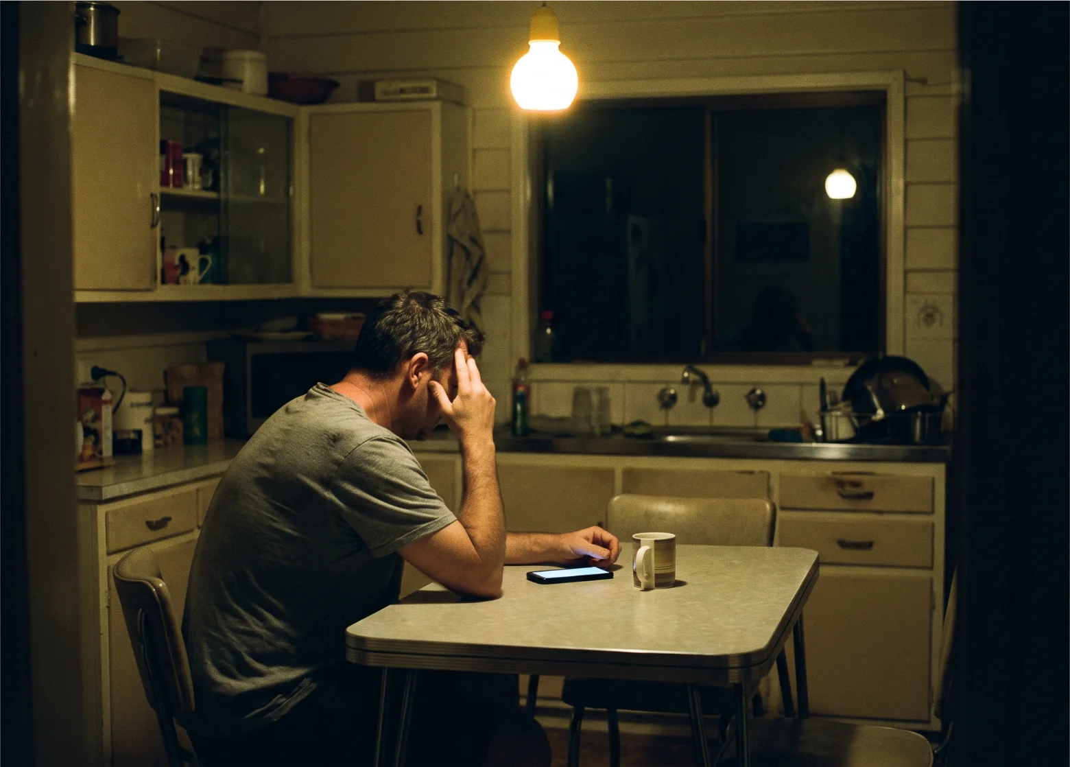 A man sits alone at a kitchen table late at night, head in one hand, a phone lying face up beside a mug.